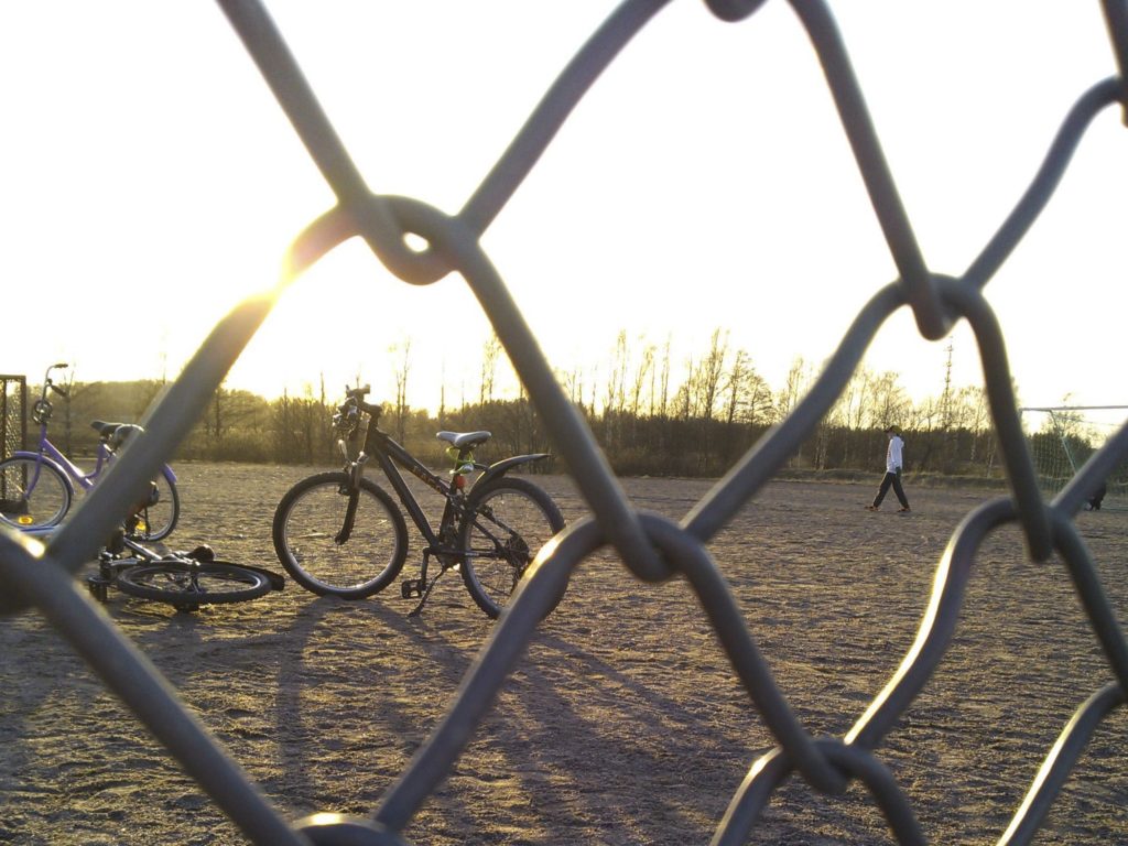 Kolme polkupyörää ja yksi kävelevä ihmishahmo kuvattuna verkkoaidan läpi laskevaa aurinkoa vasten.