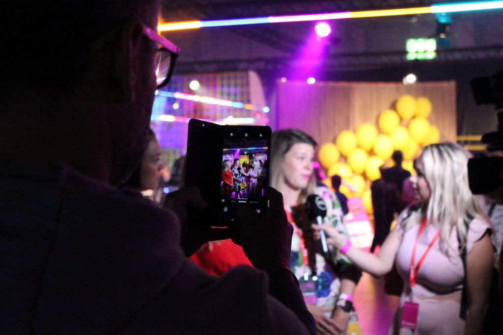 Etualalla henkilö kuvaamassa kännykkäkameralla kolmea naista, joista yksi pitää mikrofonia toisen kasvojen edessä. Taka-alalalla ihmisiä, ilmapalloja, esiintymislava ja neonvaloja.